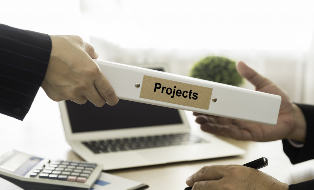 A person handing a binder labeled Projects to another business person in an office