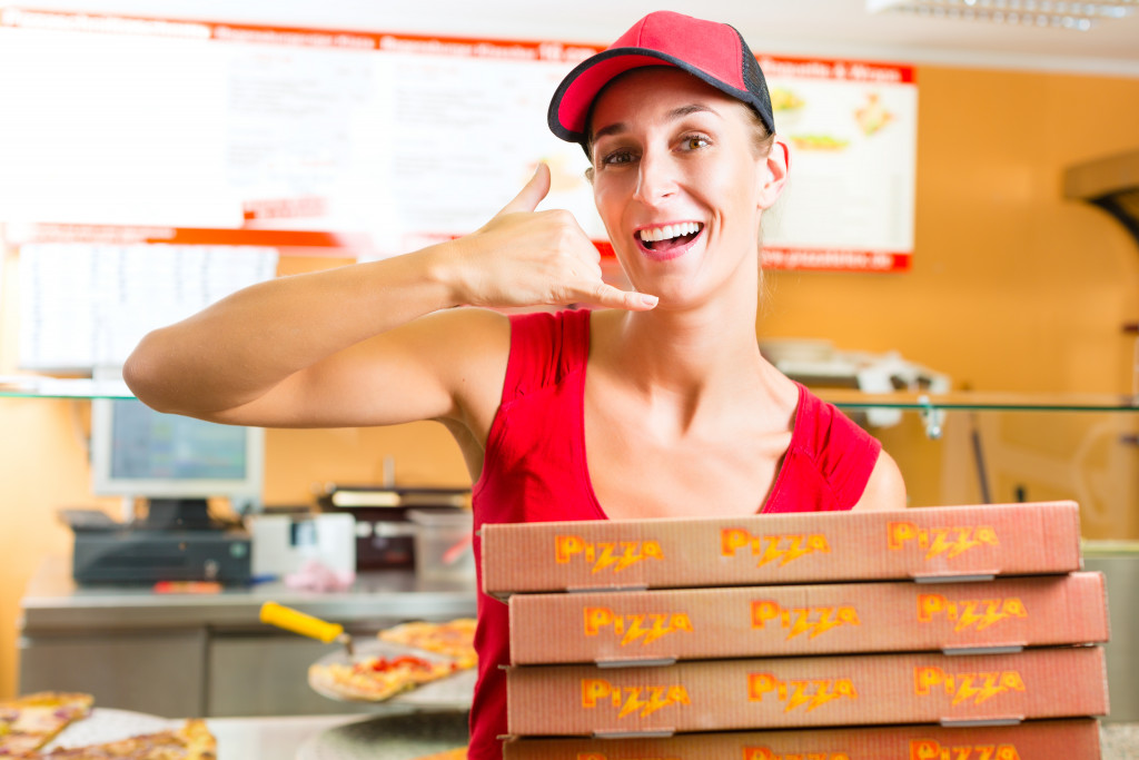 Employee smiles at camera while balancing four boxes of pizza on one hand and making a calling sign with her other hand while she is at a restaurant