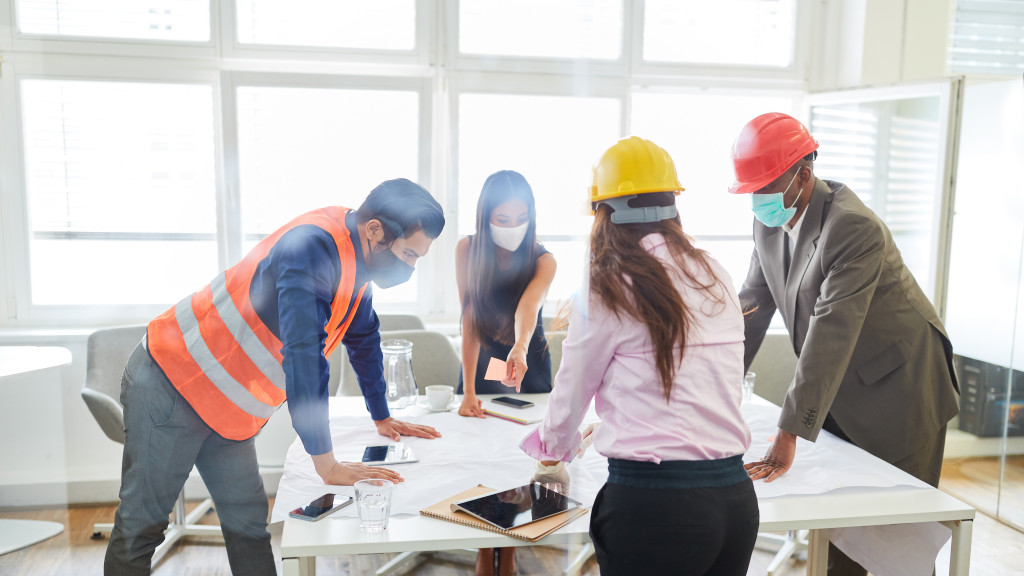 Four architects discussing the blueprint of a construction project while wearing a face mask.