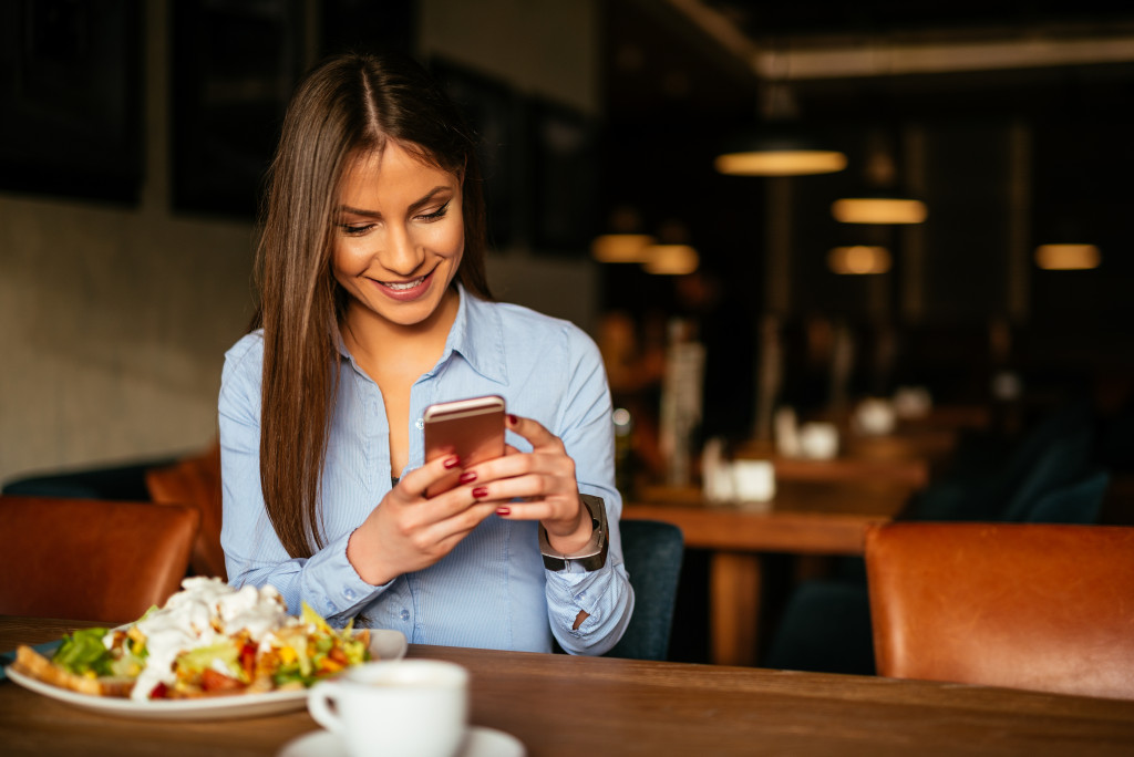 female boss smiling while using phone with healthy salad meal in front of her at the table