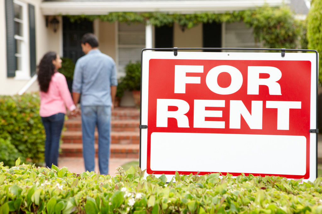 For rent sign in front of a house with a young couple checking the house.